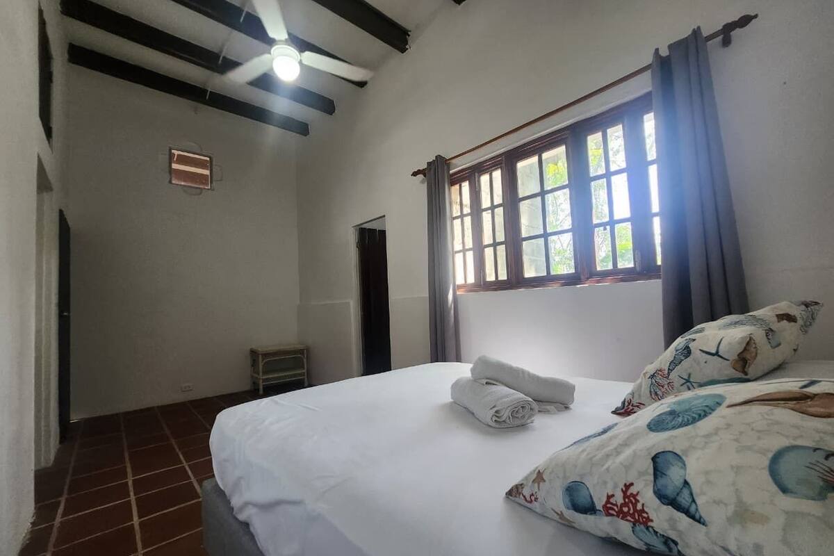 Een slaapkamer met een bed, een raam met gordijnen, een plafondventilator en een kleine tafel.