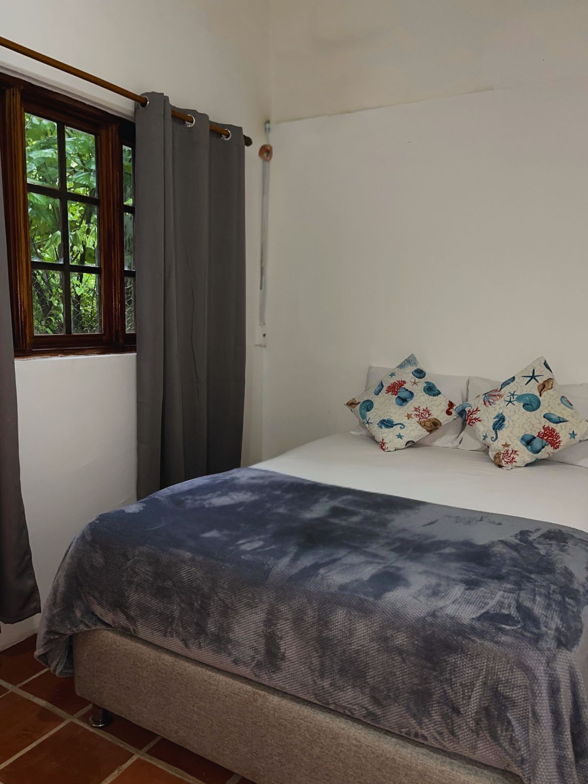Un dormitorio con cama, cortinas grises, ventana con vista a follaje y dos almohadones decorativos.