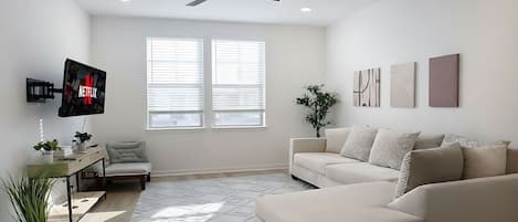 Ein modernes Wohnzimmer mit einem beigefarbenen Sofa, einem an der Wand montierten Fernseher, einem Deckenventilator und einem großen Fenster mit Jalousien.