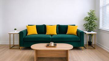 Um sofá verde com almofadas amarelas, uma mesa de centro redonda de madeira e duas mesinhas de cabeceira com plantas.