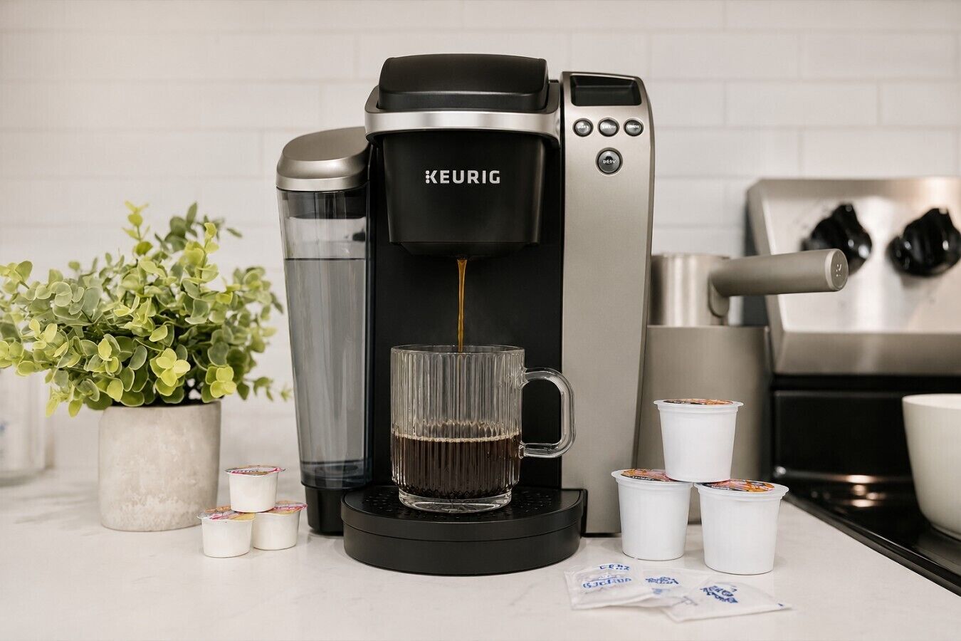 A Keurig coffee maker on a kitchen counter with a glass mug, a potted plant, and coffee pods.