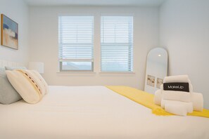 Ein Schlafzimmer mit einem Bett, einem Sessel, einem Spiegel und zwei Fenstern mit Jalousien.