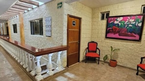 Un hall d’hôtel avec un long comptoir d’accueil, une porte en bois, un tableau au mur et deux chaises ornées de plantes en pot.