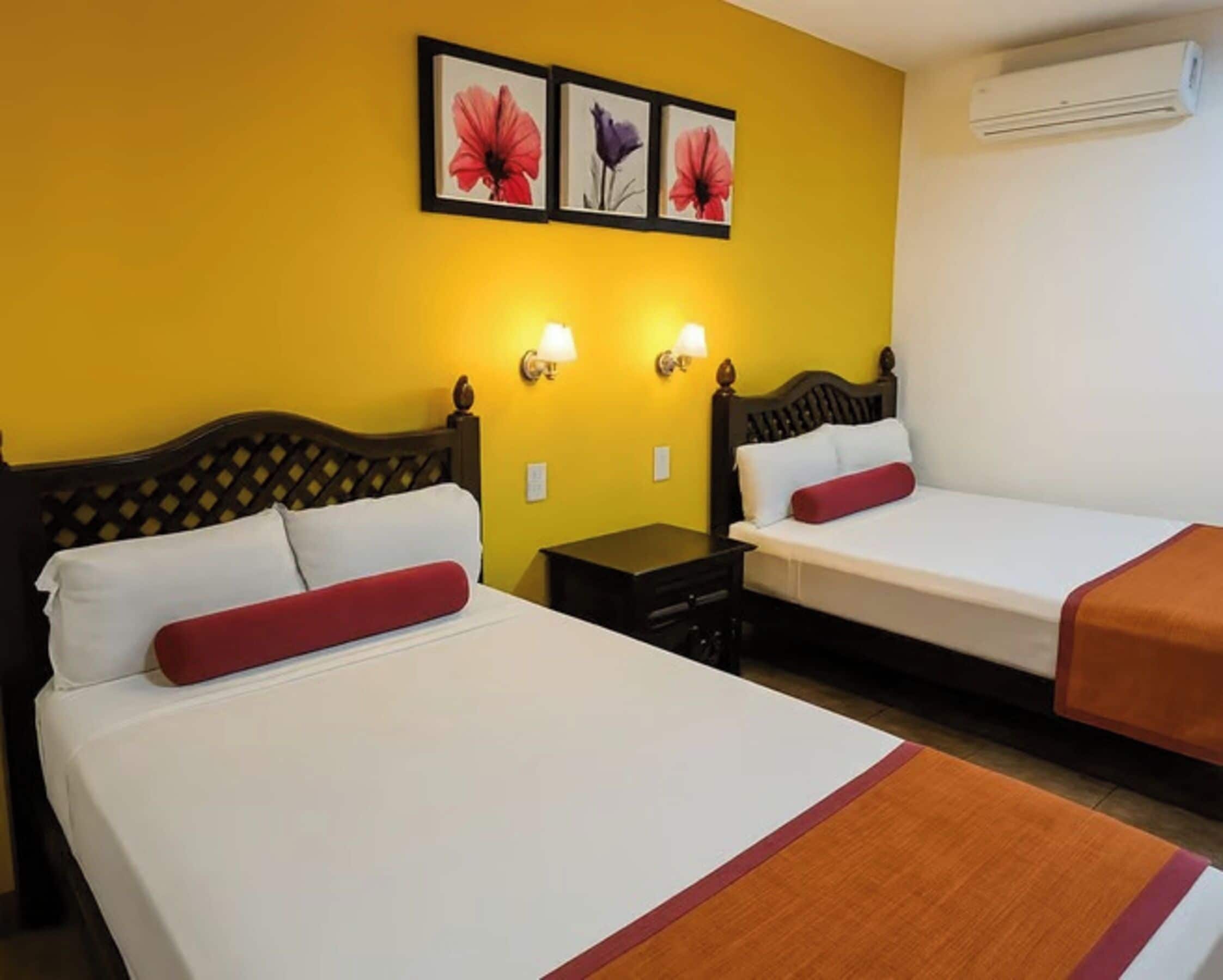Two single beds with wooden headboards, white bedding, and red pillows. There are two wall-mounted lamps, a small table, and an air conditioning unit.