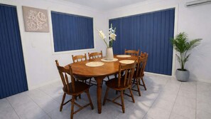 A dining room with a round wooden table, six wooden chairs, a vase with flowers, and a potted plant. - Pebbly Beach Escape | Sea View (Forster)