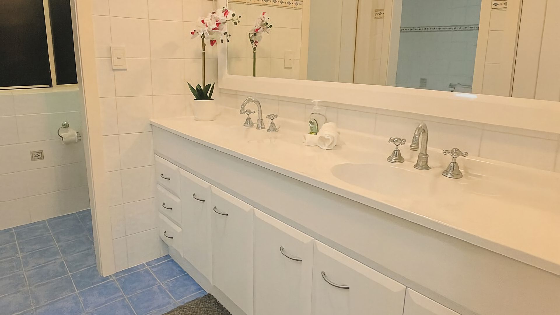 Ein Badezimmer mit einem weißen Waschtisch, einem Spiegel, einem Waschbecken mit zwei Wasserhähnen und einer Orchidee im Topf.