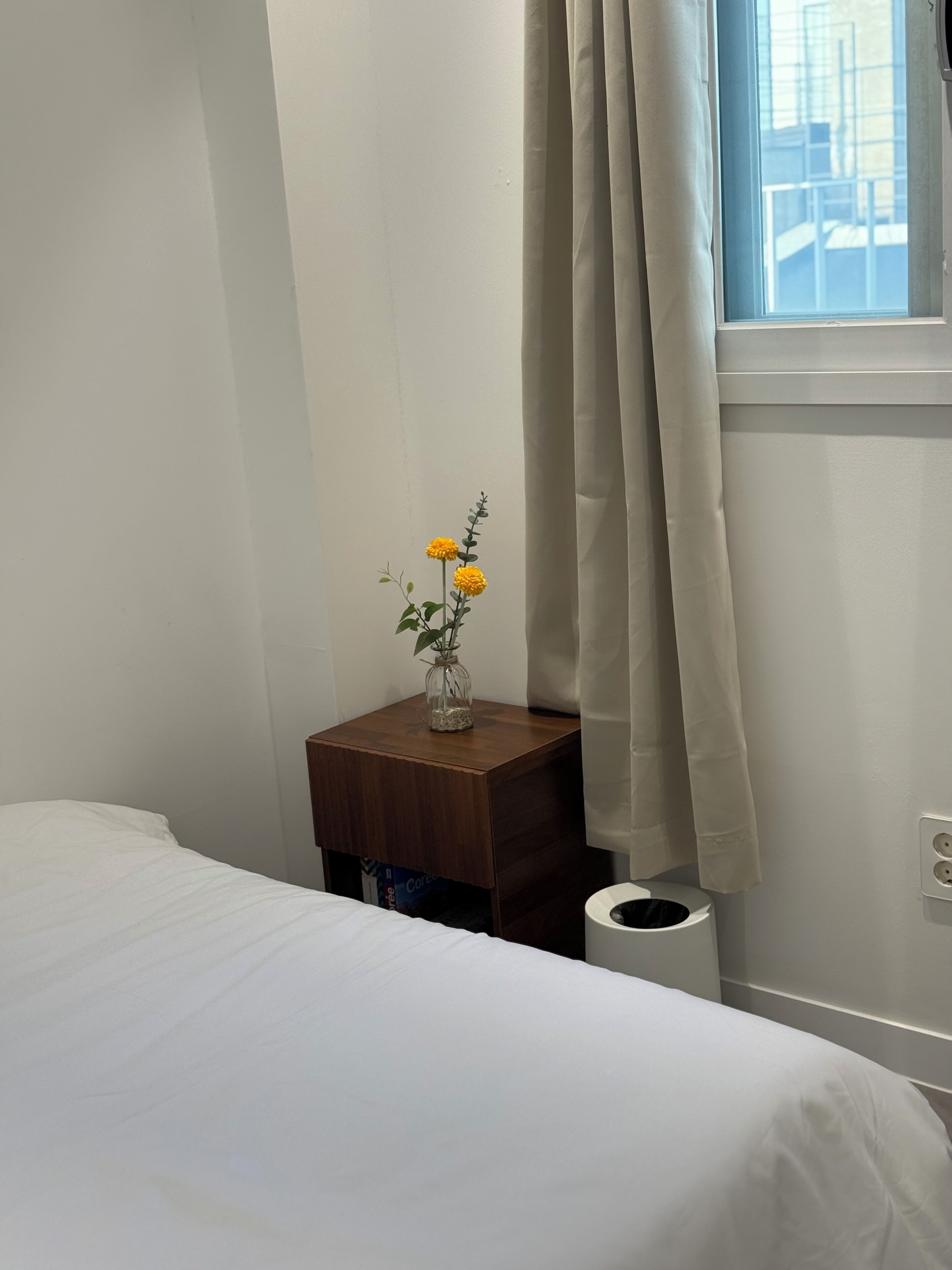 Een slaapkamer met een bed, een nachtkastje met een bloemenvaas, een raam met gordijnen en een stopcontact.