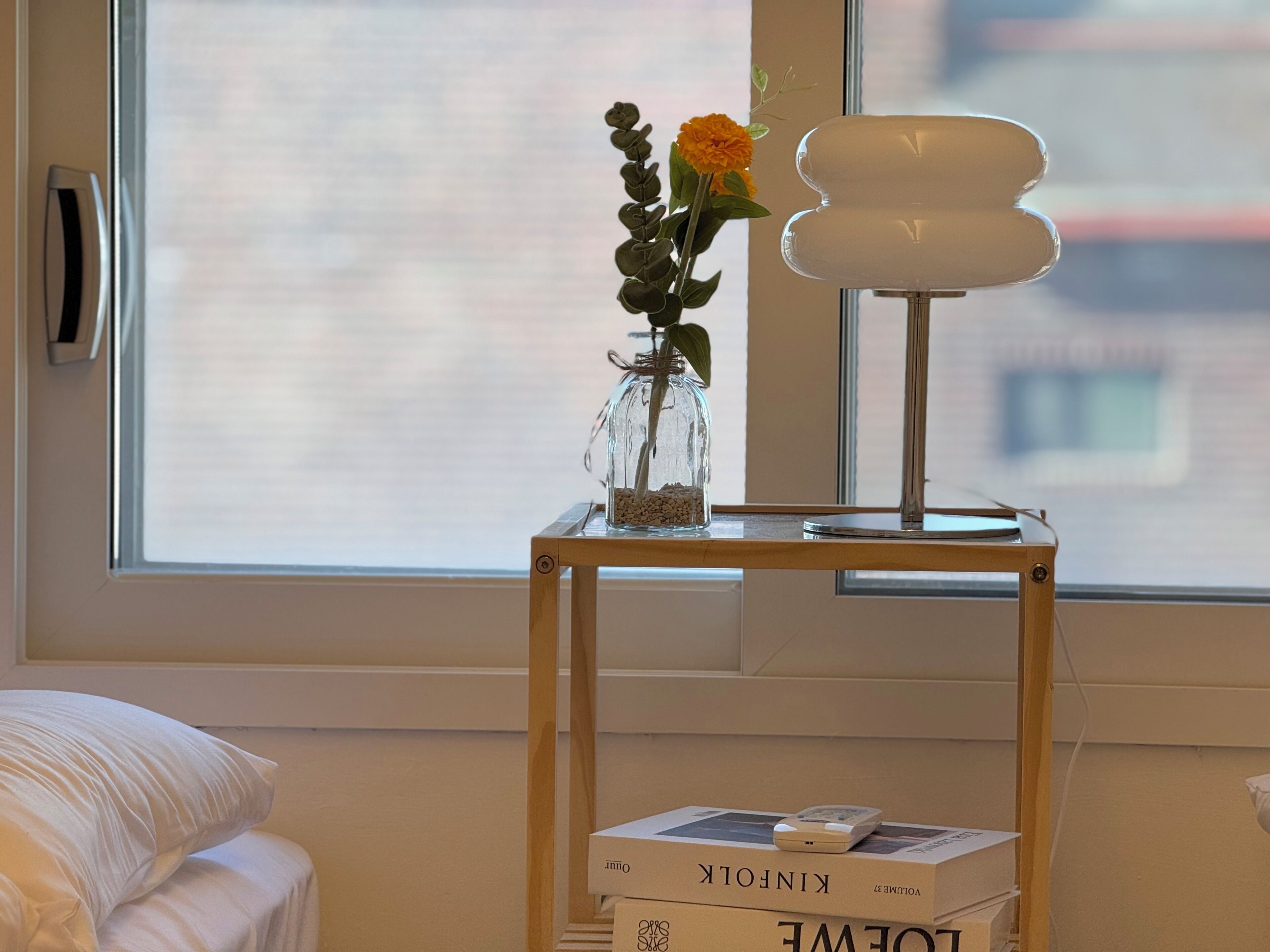 A room with a window, a bedside table with a lamp, a vase with flowers, and books.