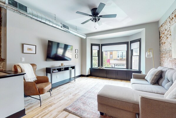 Una sala de estar moderna con una televisión de pantalla plana, un ventilador de techo, un sofá, una silla y un ventanal con vistas a un edificio.
