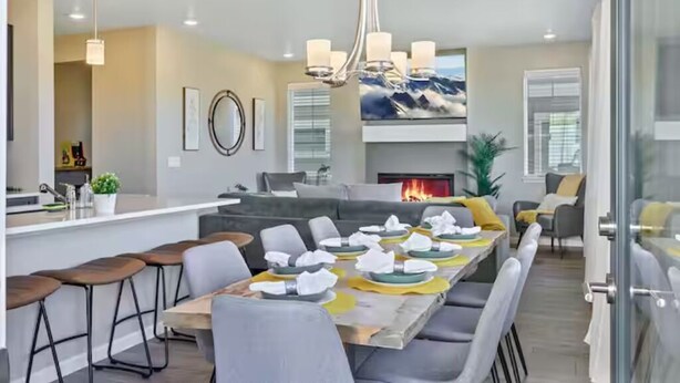 A modern dining area with a long table set for a meal, a kitchen area with bar stools, and a fireplace. - Family Friendly 5BR Mountain Views & Game Room with AC (Superior)