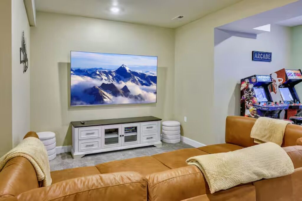 Una sala de estar con un televisor de pantalla plana grande montado en la pared, un sofá de cuero marrón, un mueble blanco para entretenimiento y un letrero en la pared que dice "ARCADE".