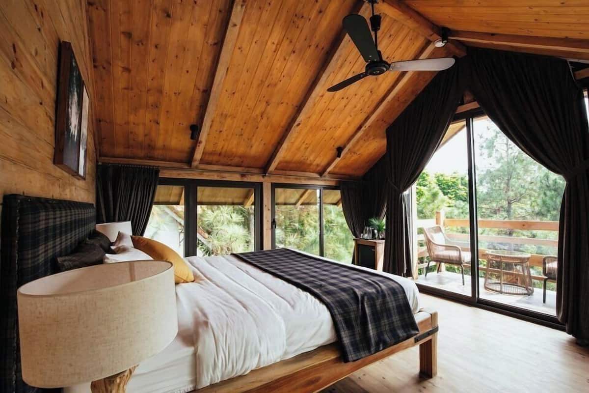 Eine Holzbalkendecke mit Deckenventilator, ein Bett mit Kopfkissen und Decke, ein Tisch mit Stühlen und Blick auf Grünlandschaft im Freien.