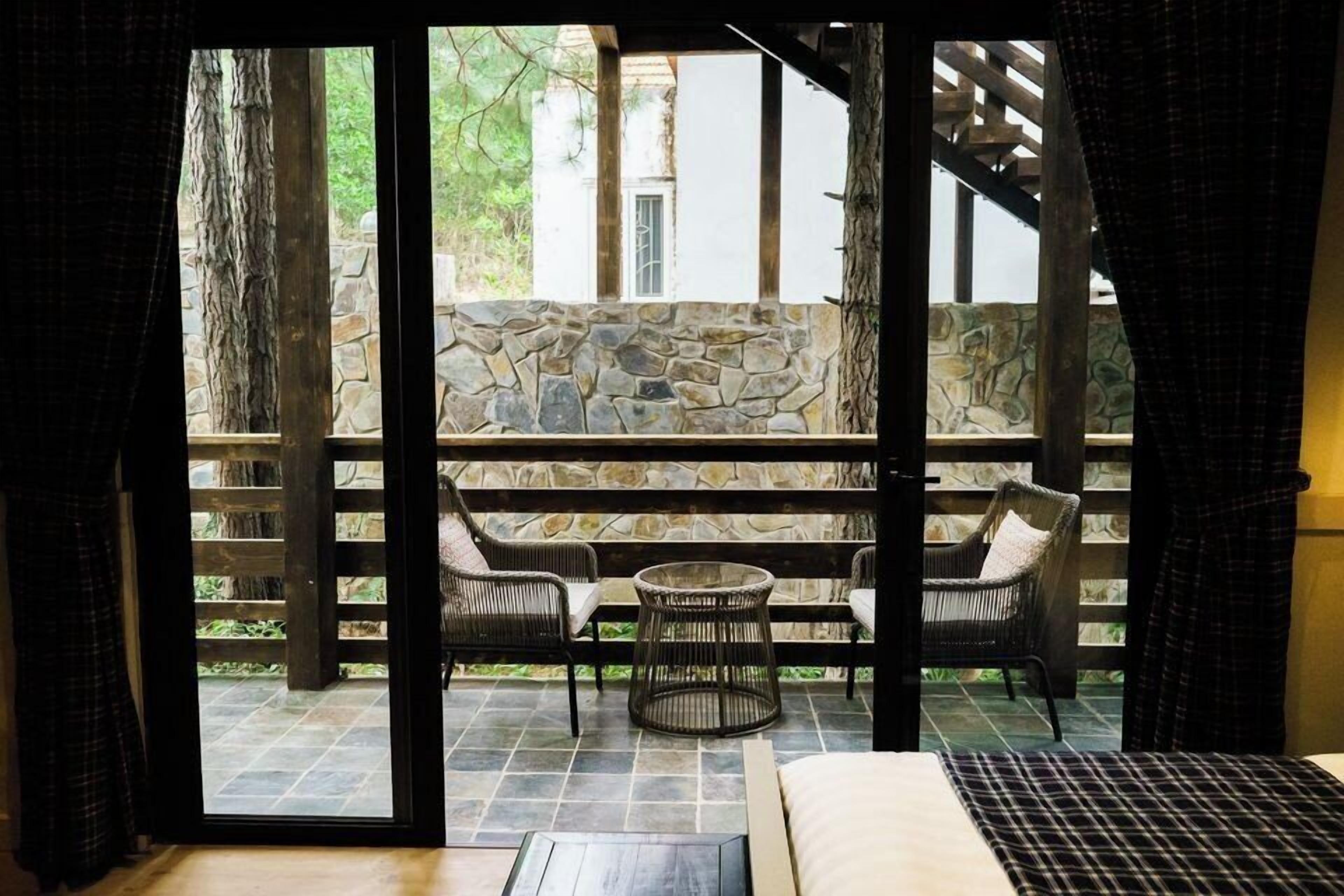 A room with a stone wall, a wooden ceiling, a bed, and a window with a view of a stone wall and greenery outside.