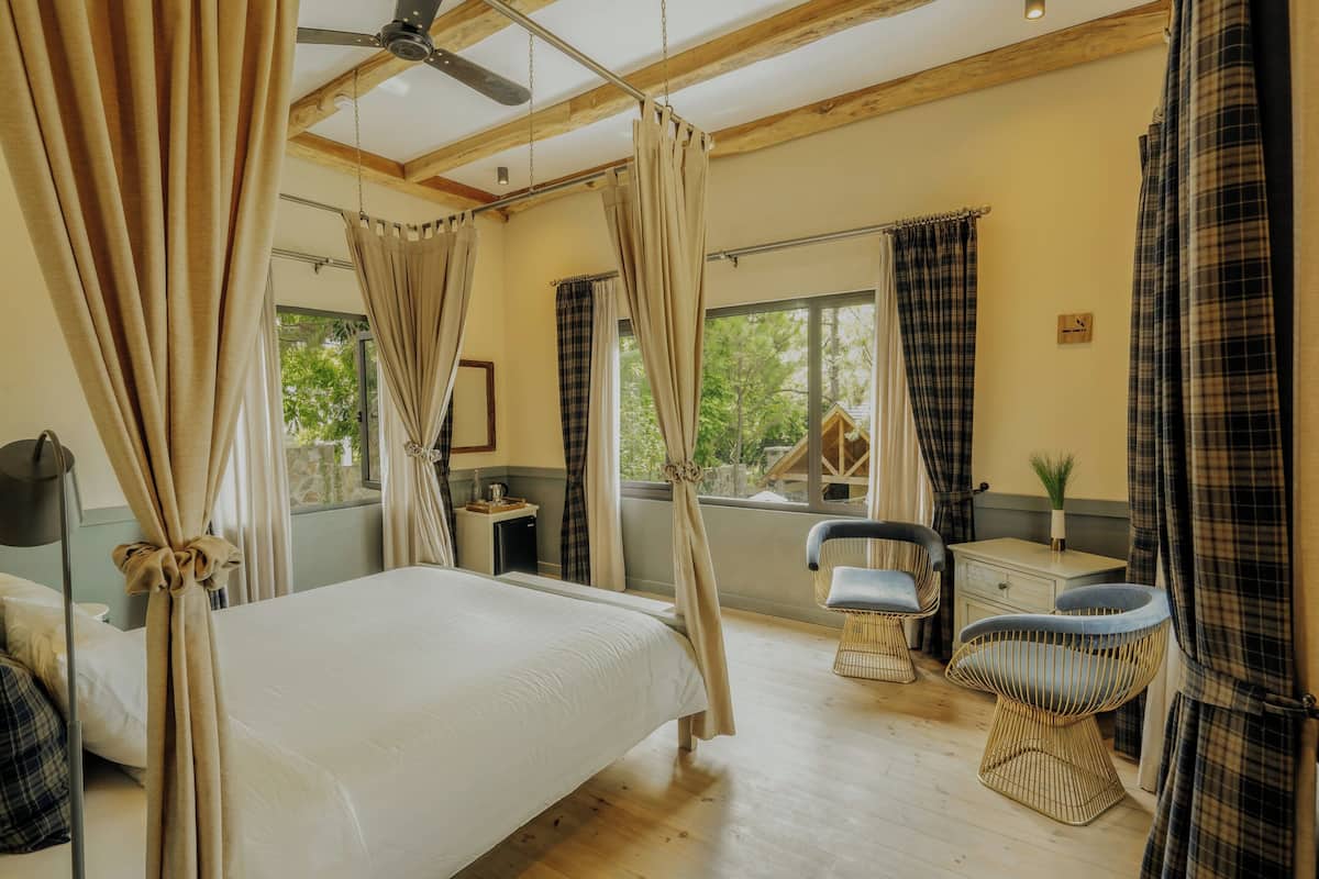Ein Schlafzimmer mit Bett, Deckenventilator, Fenster mit Vorhängen, einem Sessel und einem kleinen Tisch.