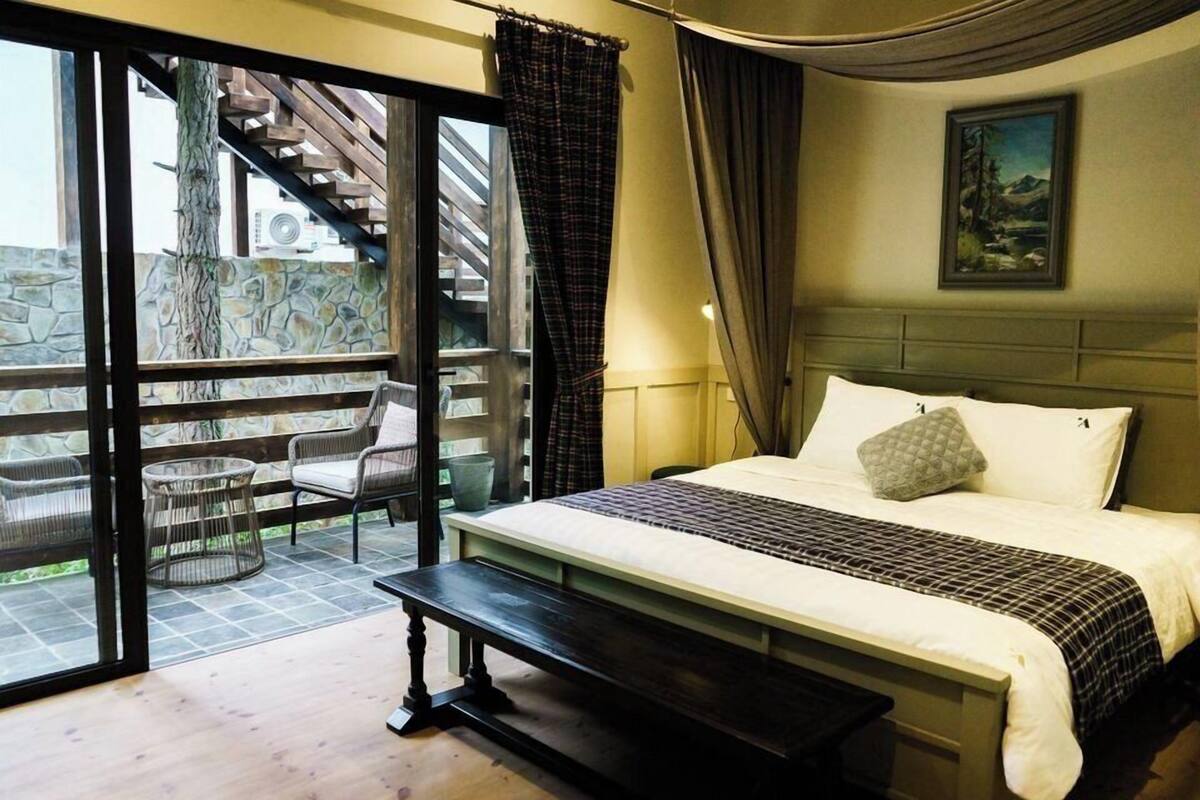 Ein Schlafzimmer mit einem Bett, einer Bank, einem Sessel und Blick auf einen Balkon mit einer Steinmauer und einer Holztreppe.