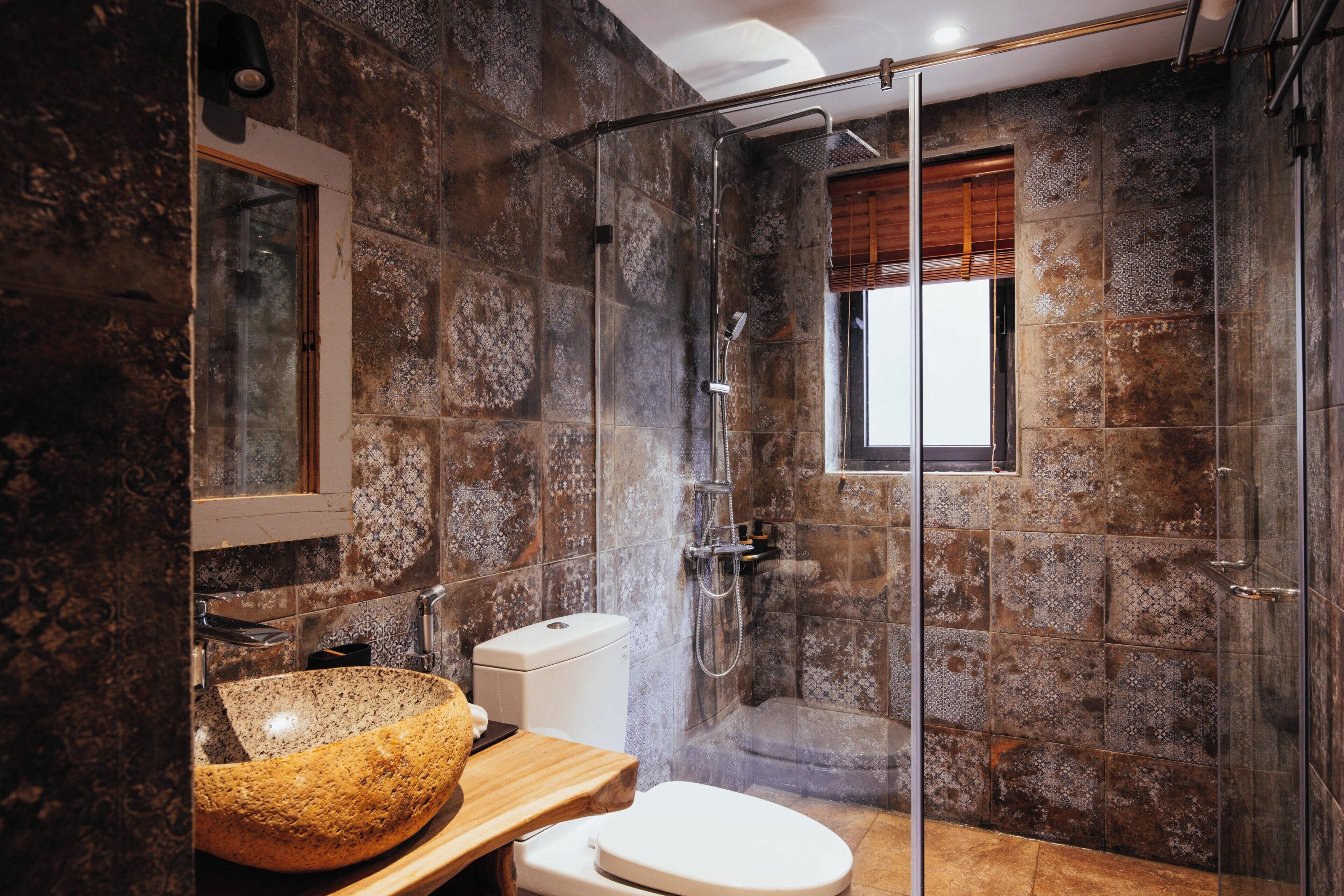 Un baño con pared de piedra, ducha envuelta en vidrio, tocador de madera y un inodoro blanco.
