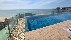 A clear view of a swimming pool with a wooden deck and a glass barrier, overlooking a beach and the sea. - Avatara Beachfront - Best Rooftop Pool & Ocean Views (Pattaya)