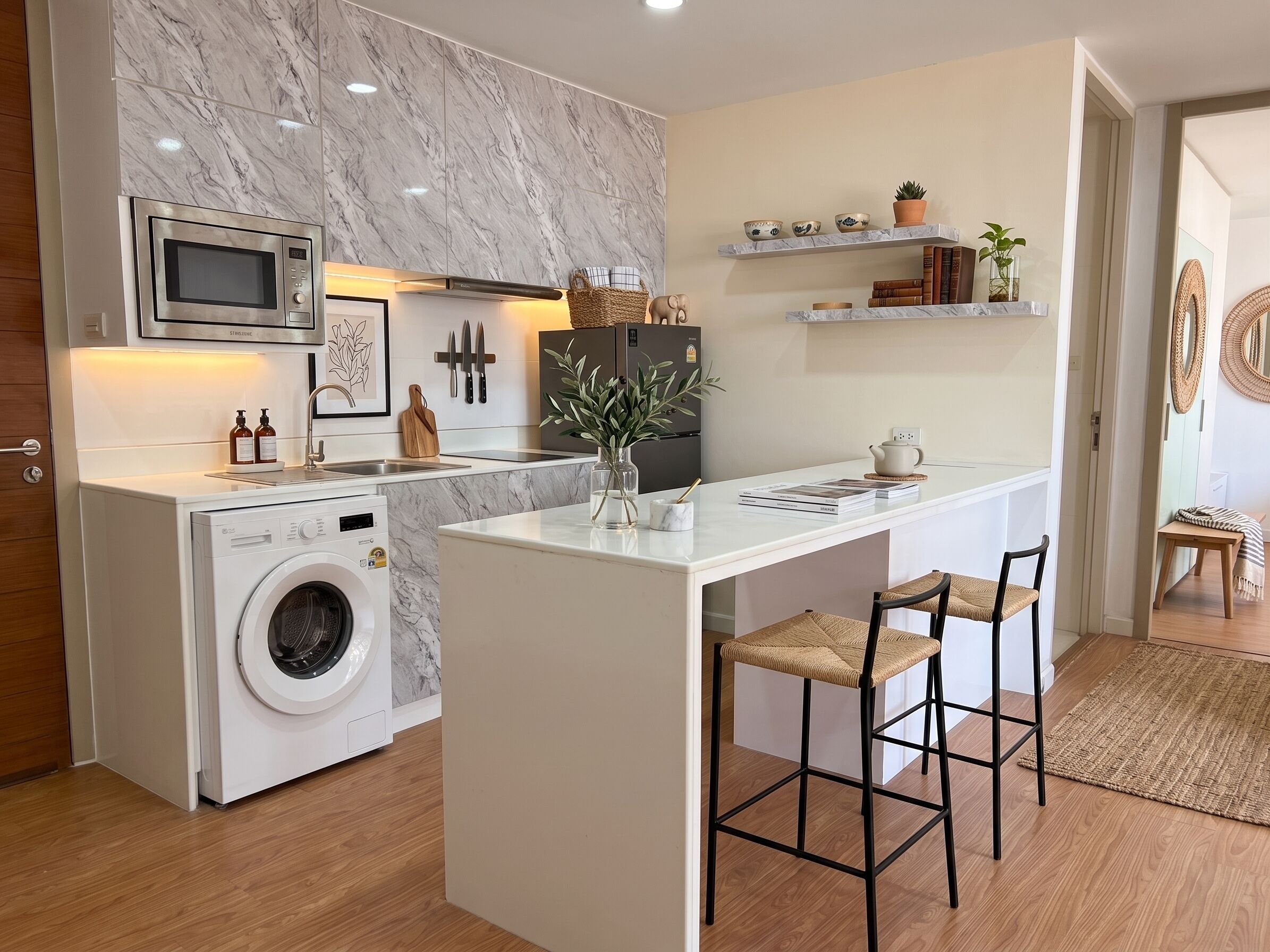 Cocina compacta con lavadora integrada, microondas y refrigerador. Hay una pequeña isla con taburetes de bar y un florero con follaje.