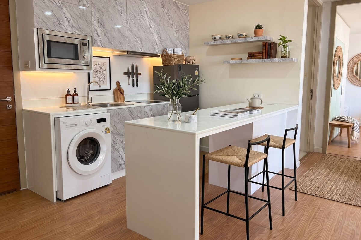 Une cuisine compacte équipée d’un lave-linge, d’un four à micro-ondes et d’un réfrigérateur. On y trouve également un petit îlot avec des tabourets de bar et un vase agrémenté de plantes.