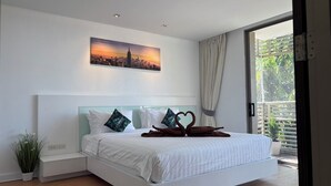 A modern bedroom with a large bed, a cityscape painting, a potted plant, and a view of greenery through a window. - Avatara Beachfront - Best Rooftop Pool & Ocean Views (Pattaya)