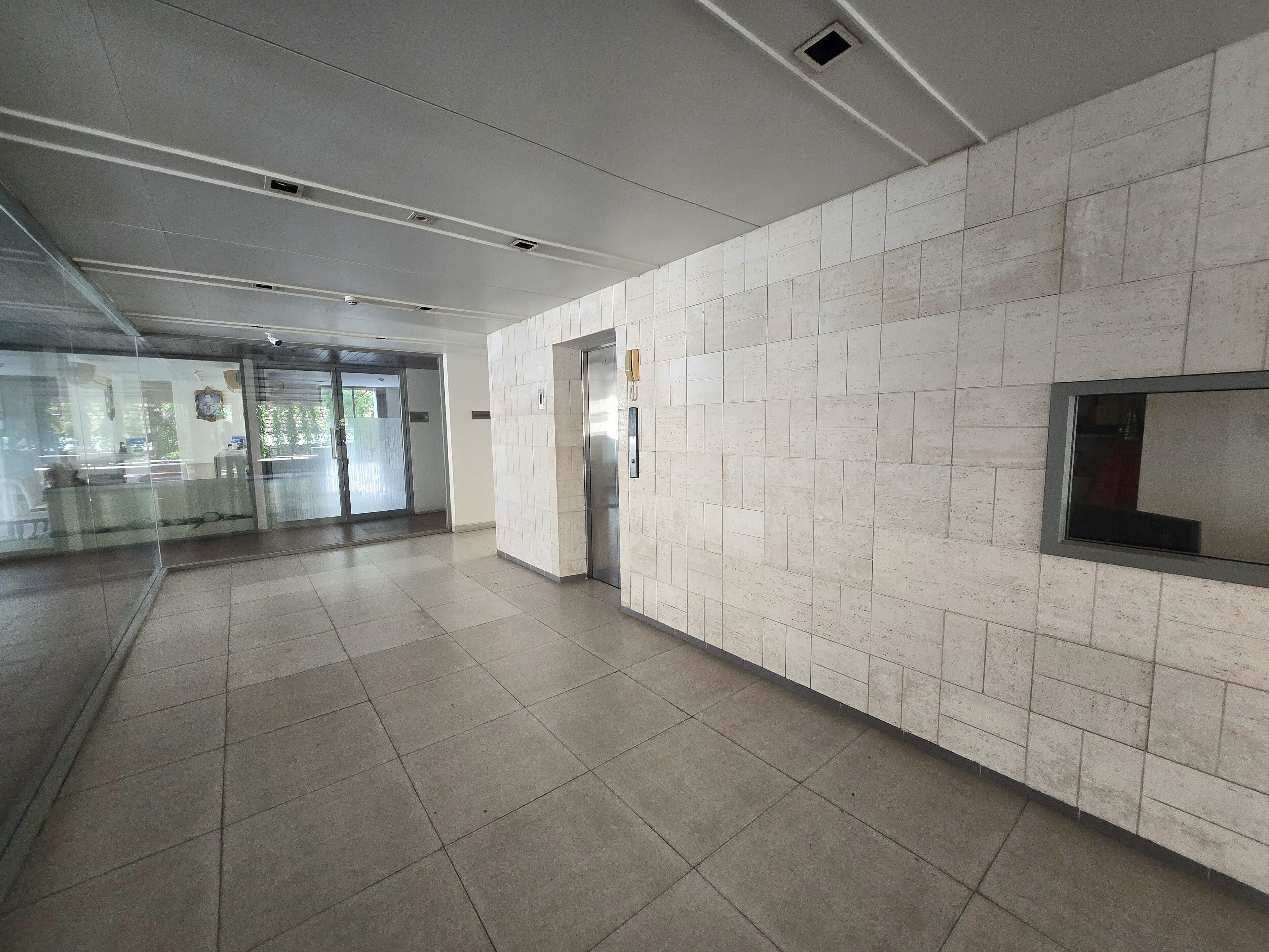 A spacious corridor with tiled walls and a tiled floor, leading to an elevator and a glass door entrance.
