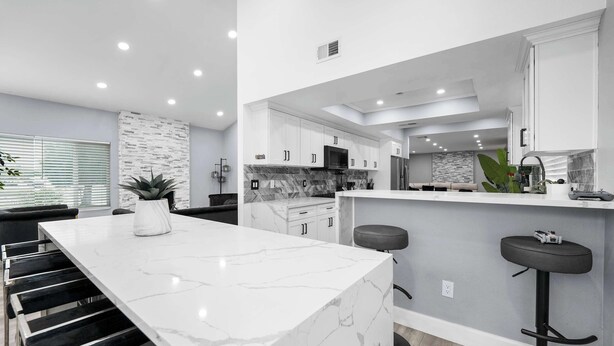 A modern kitchen with a marble island, white cabinets, and a black bar stool. - Glendora Super House! (Glendora)