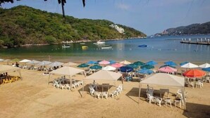 A beach with umbrellas and chairs, boats on the water, and a hill with buildings.