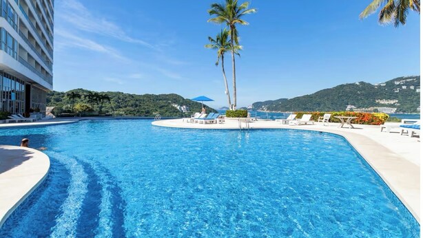 A large swimming pool with lounge chairs and palm trees, surrounded by a modern building and a scenic hillside view.