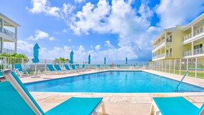 Una piscina con lettini e ombrelloni, circondata da una recinzione, con vista diretta sull'oceano e un edificio sullo sfondo.