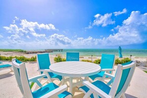 Ein Sitzbereich am Strand mit einem runden Tisch und blauen Stühlen, mit Blick auf das Meer und einen Steg.
