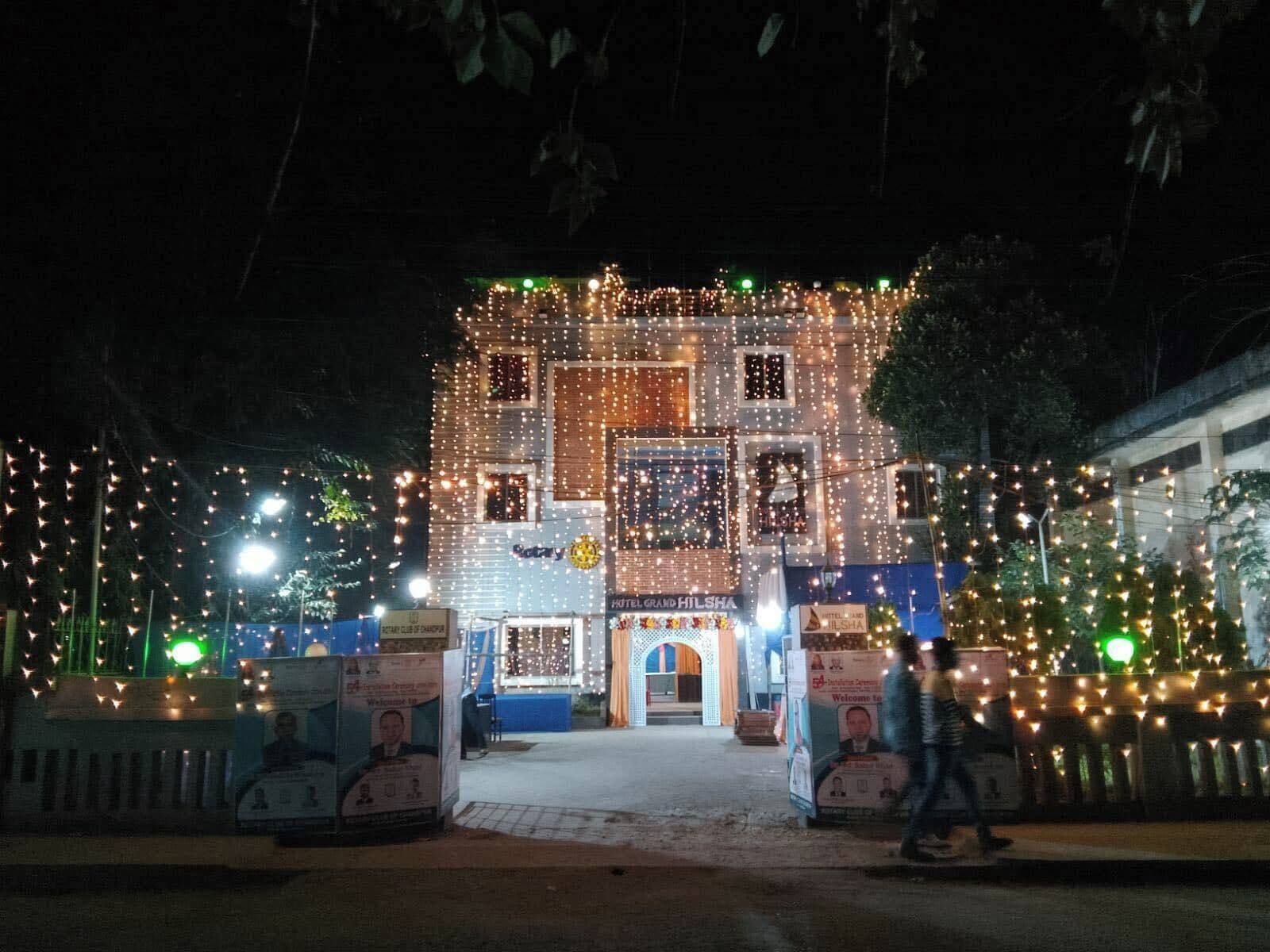 A hotel facade adorned with numerous lights, featuring a central entrance with a sign for 'Hotel Grand Hilsha'.