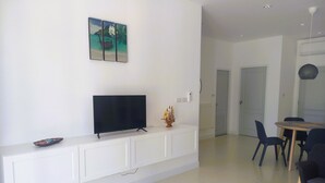 Ett modernt vardagsrum med en platt-TV på ett vitt skåp, en matplats med bord och stolar samt en hall som leder till ett annat rum.