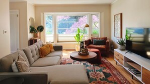 Ein Wohnzimmer mit einer Couch, einem Sessel, einem Couchtisch, einer TV-Kommode und großen Fenstern mit Blick auf Bäume.