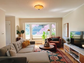 Sala de estar con sofá, sillón, mesa de centro, mueble para televisión y ventanas amplias con vistas a árboles.