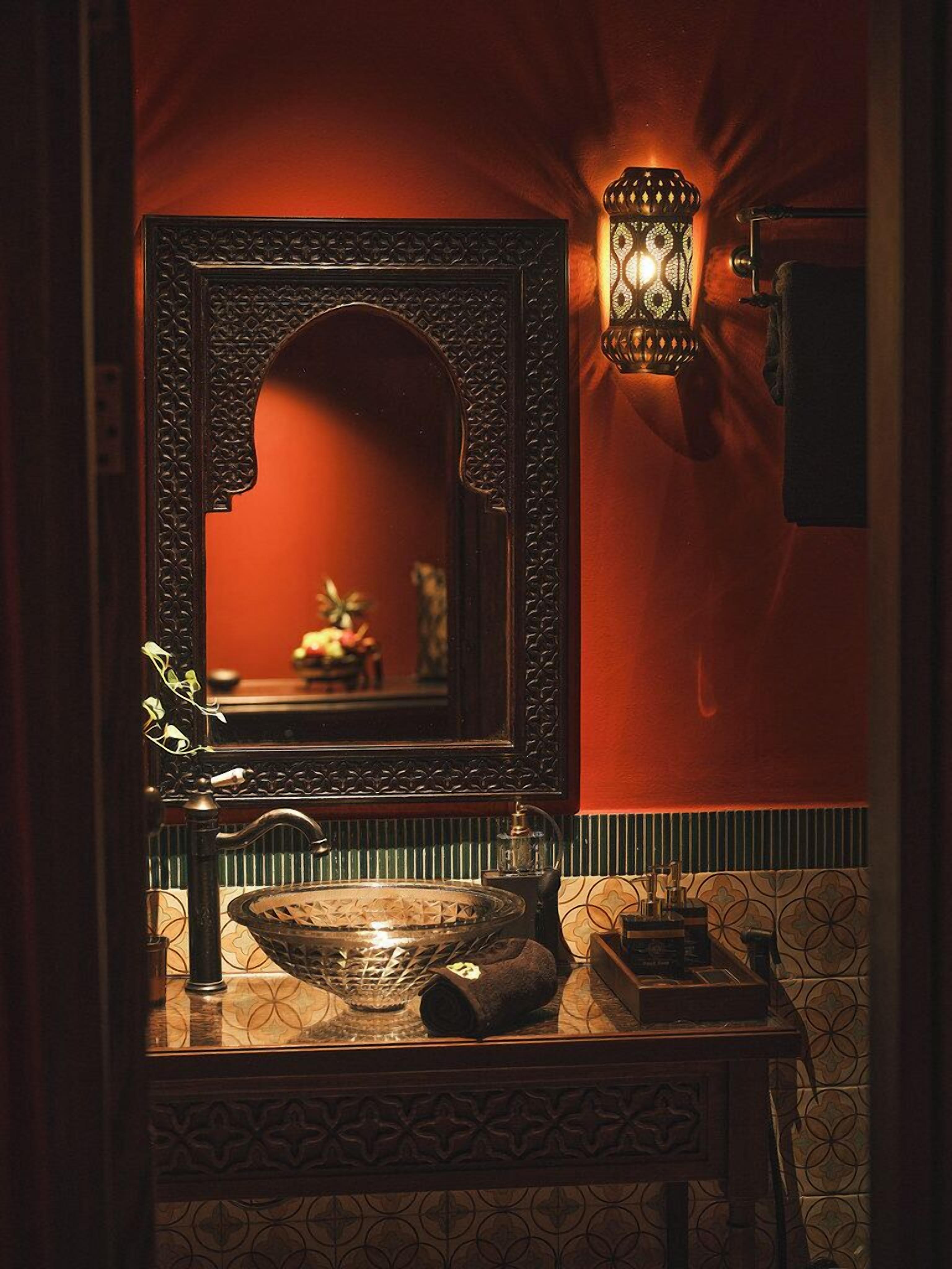 A bathroom with a large ornate mirror, a wall-mounted light fixture, a sink with a glass bowl, and patterned tiles.