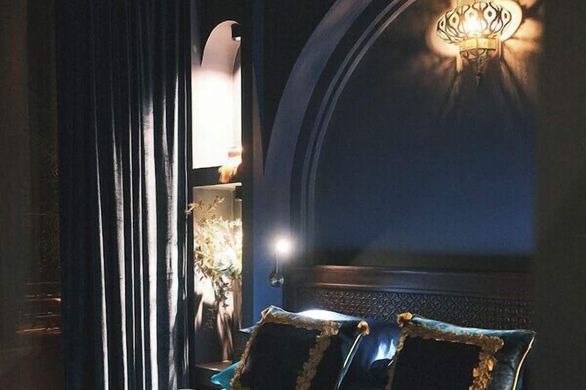A bed with dark blue bedding and gold trim, a wall-mounted lamp, and a dark curtain.