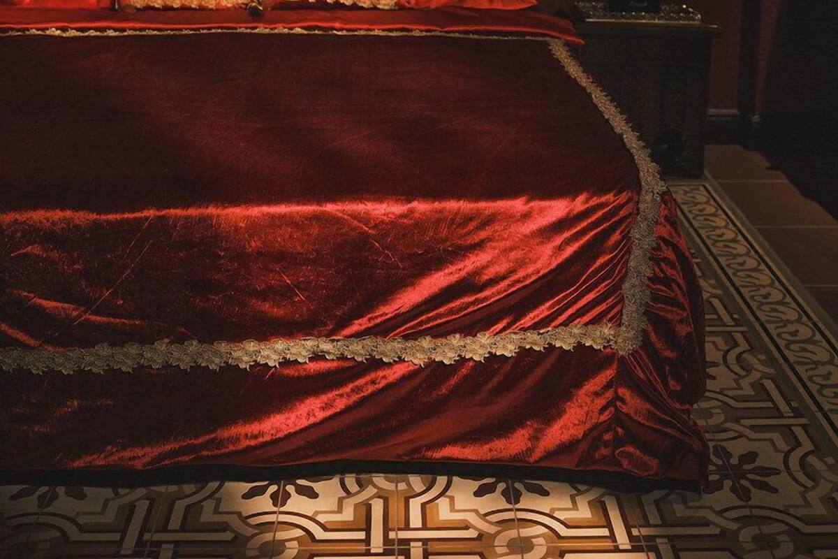 A bed with a red velvet bedspread and matching pillows, two wall-mounted lamps, and a patterned floor.