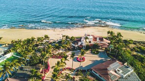 Vista aérea de un complejo turístico costero con playa, palmeras y un edificio de techo rojo.