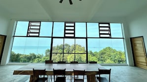 A spacious room with a large table and chairs, large windows with a view of the sea, and a ceiling fan. - Ferringhi Inn 51 (George Town)