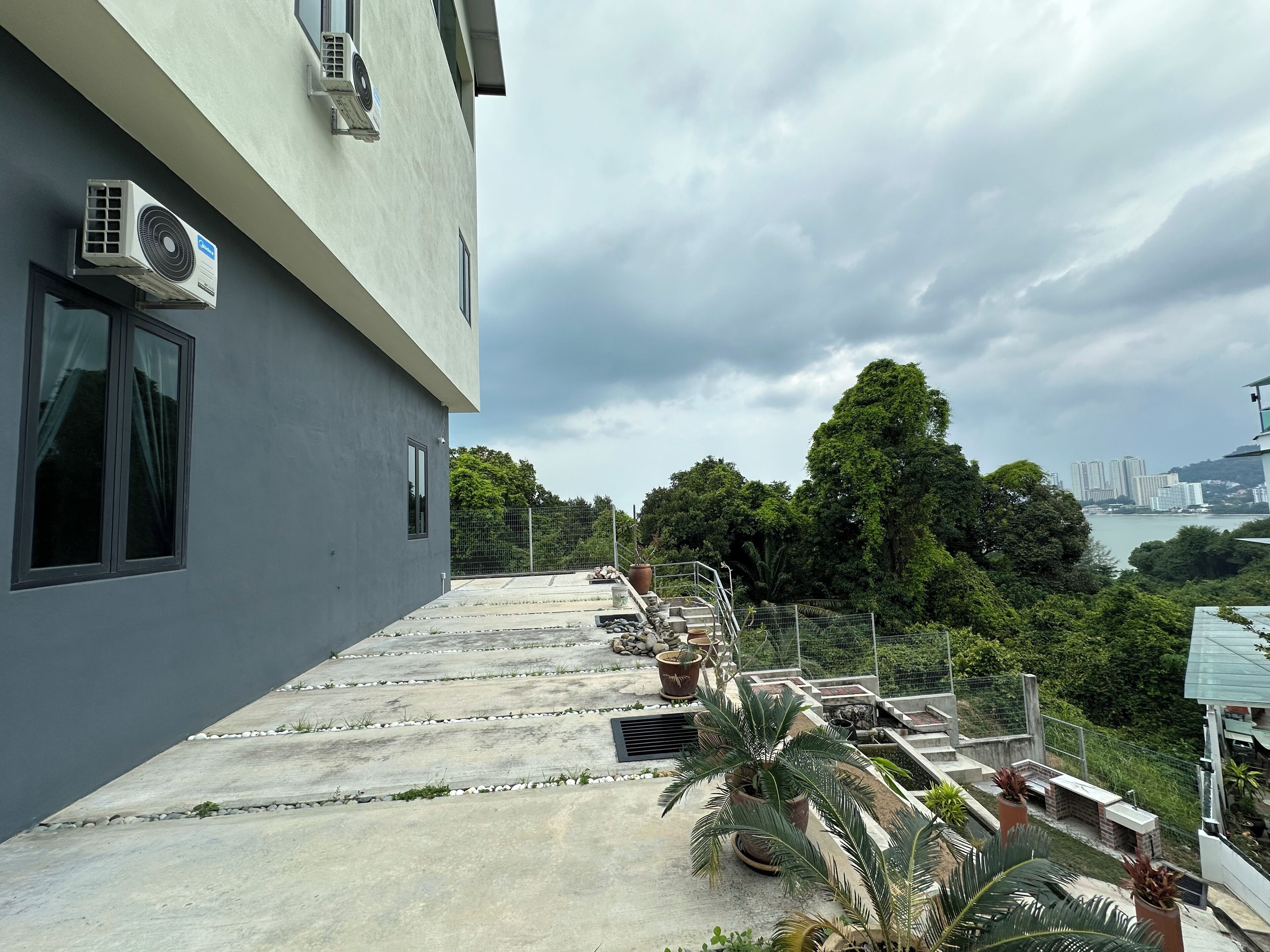 A rooftop terrace with potted plants, a view of a cityscape, and a building with air conditioning units.