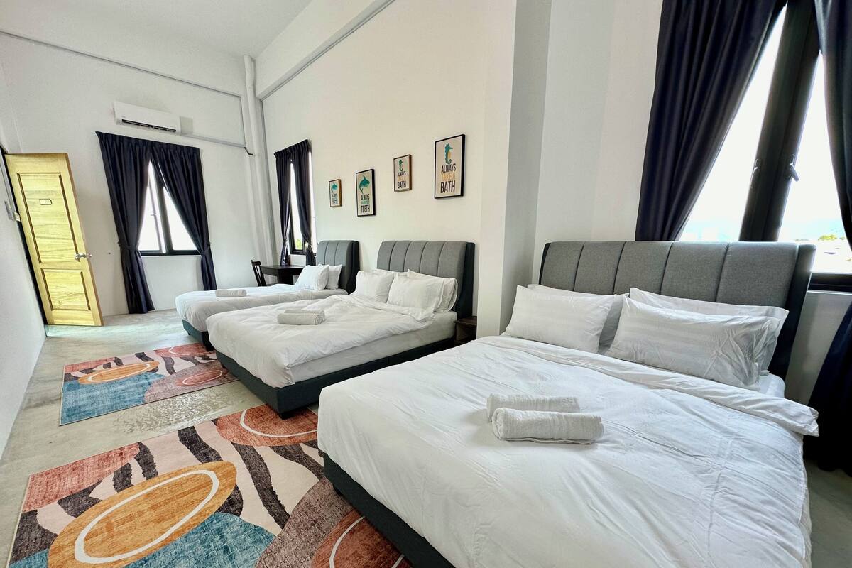 A hotel room with two single beds, a large bed, a desk, and a chair. There are air conditioning units on the wall, a window with curtains, and a door.