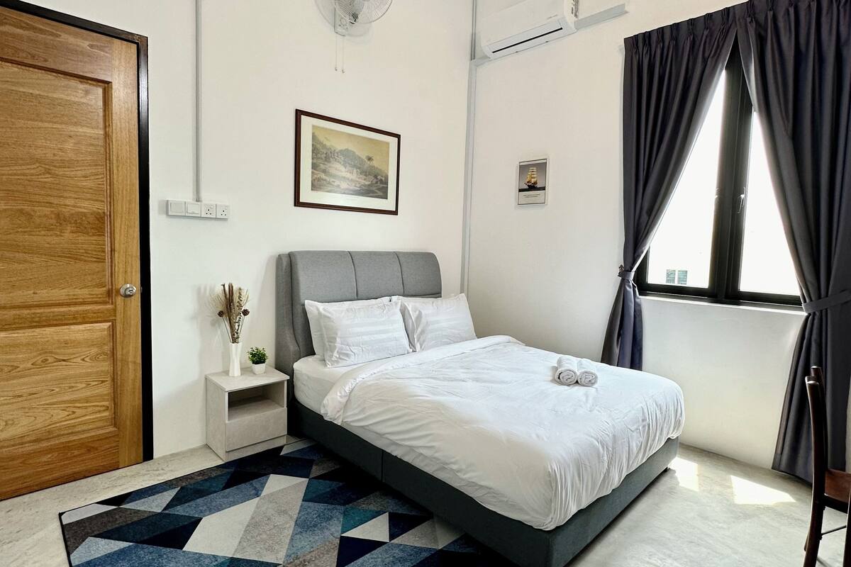 A modern bedroom with a bed, a bedside table, a wall-mounted fan, a window with curtains, and a geometric-patterned rug.