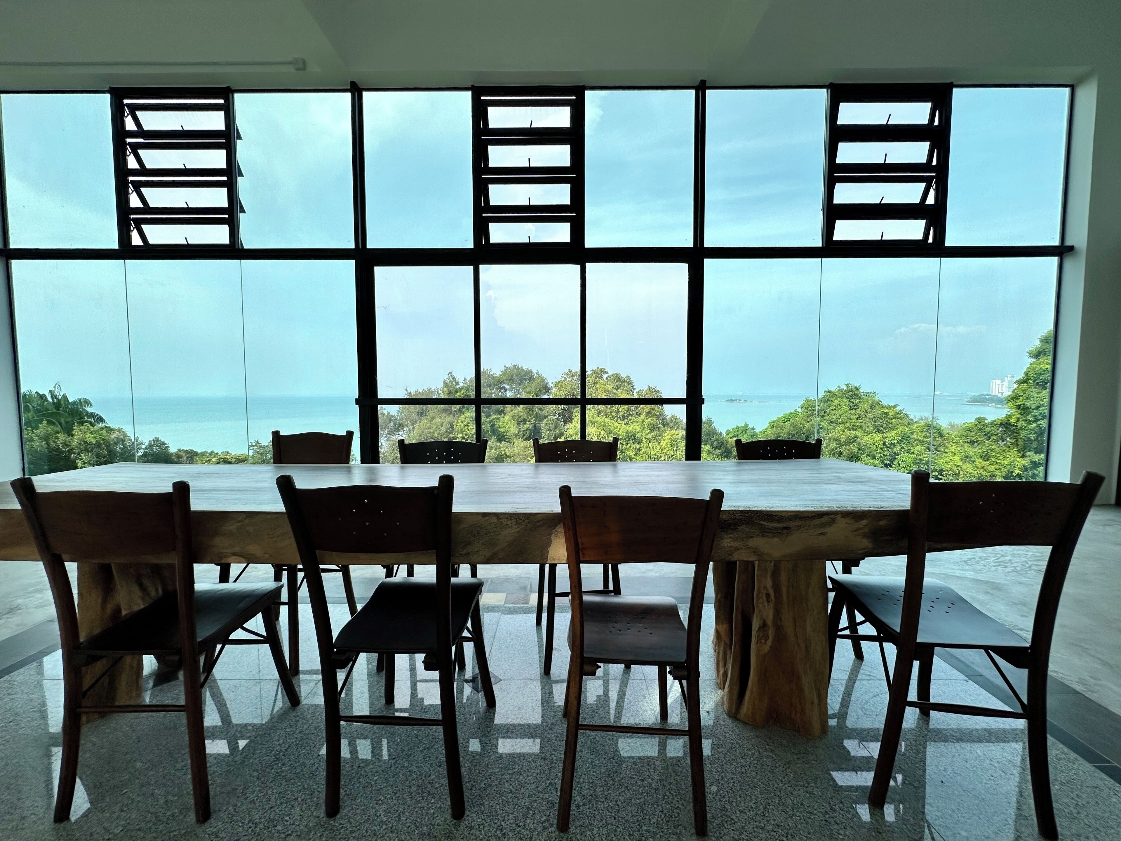 A large dining table with six chairs in a modern room with large windows offering a view of the sea and greenery.