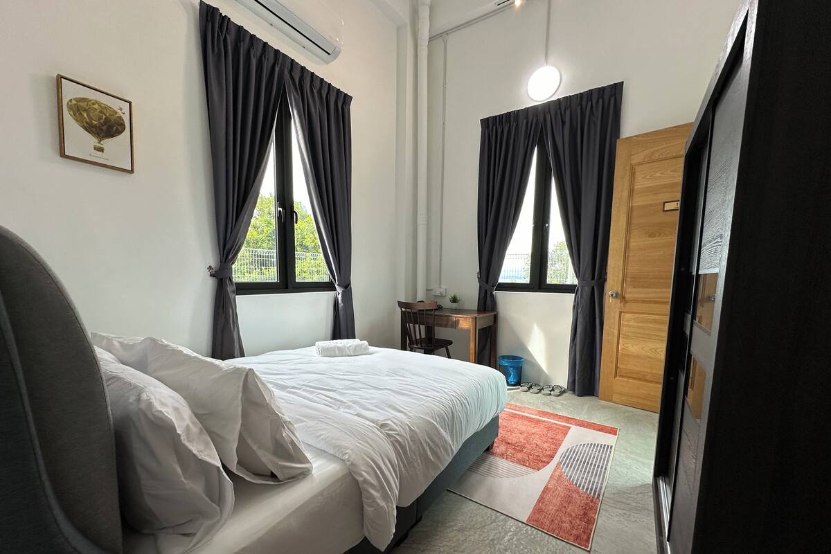 A bedroom with a bed, a desk, and two windows with curtains.
