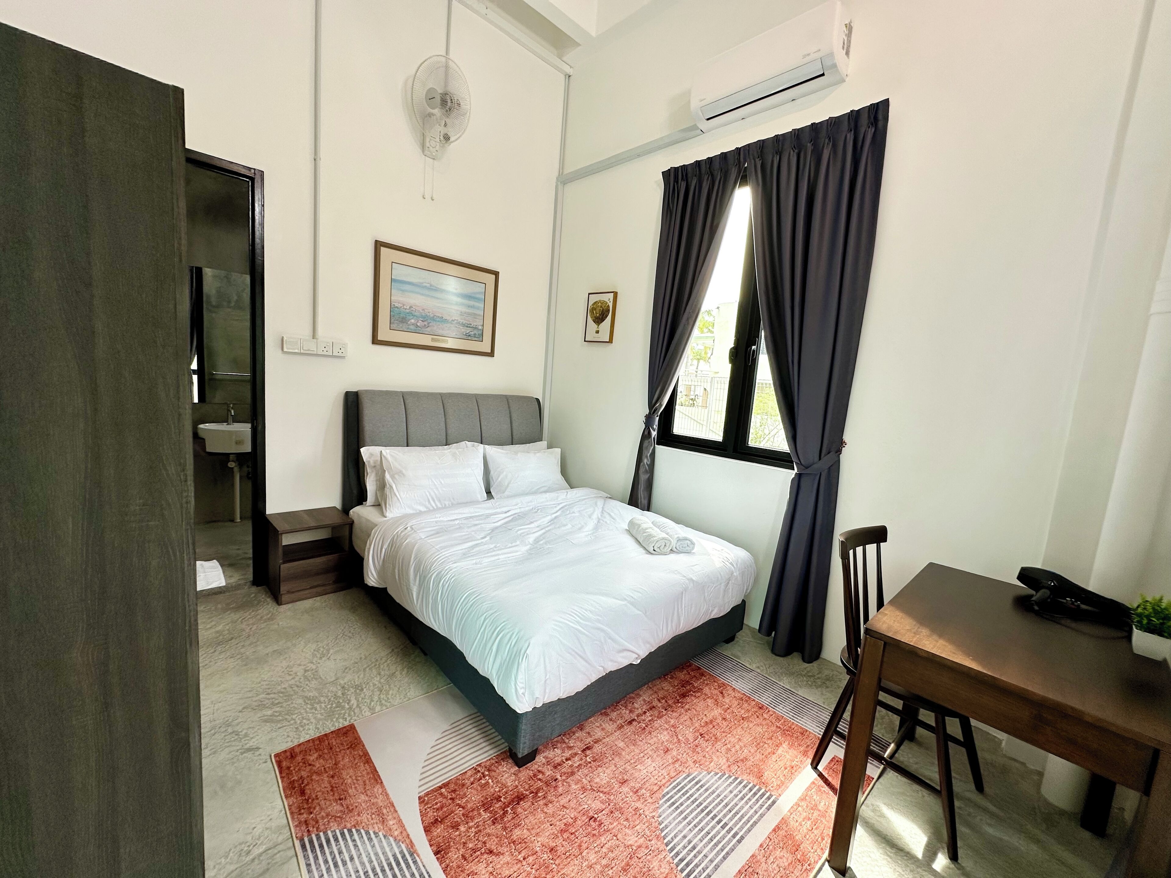 A bedroom with a bed, a desk, a chair, a window with curtains, a picture on the wall, and an air conditioner.