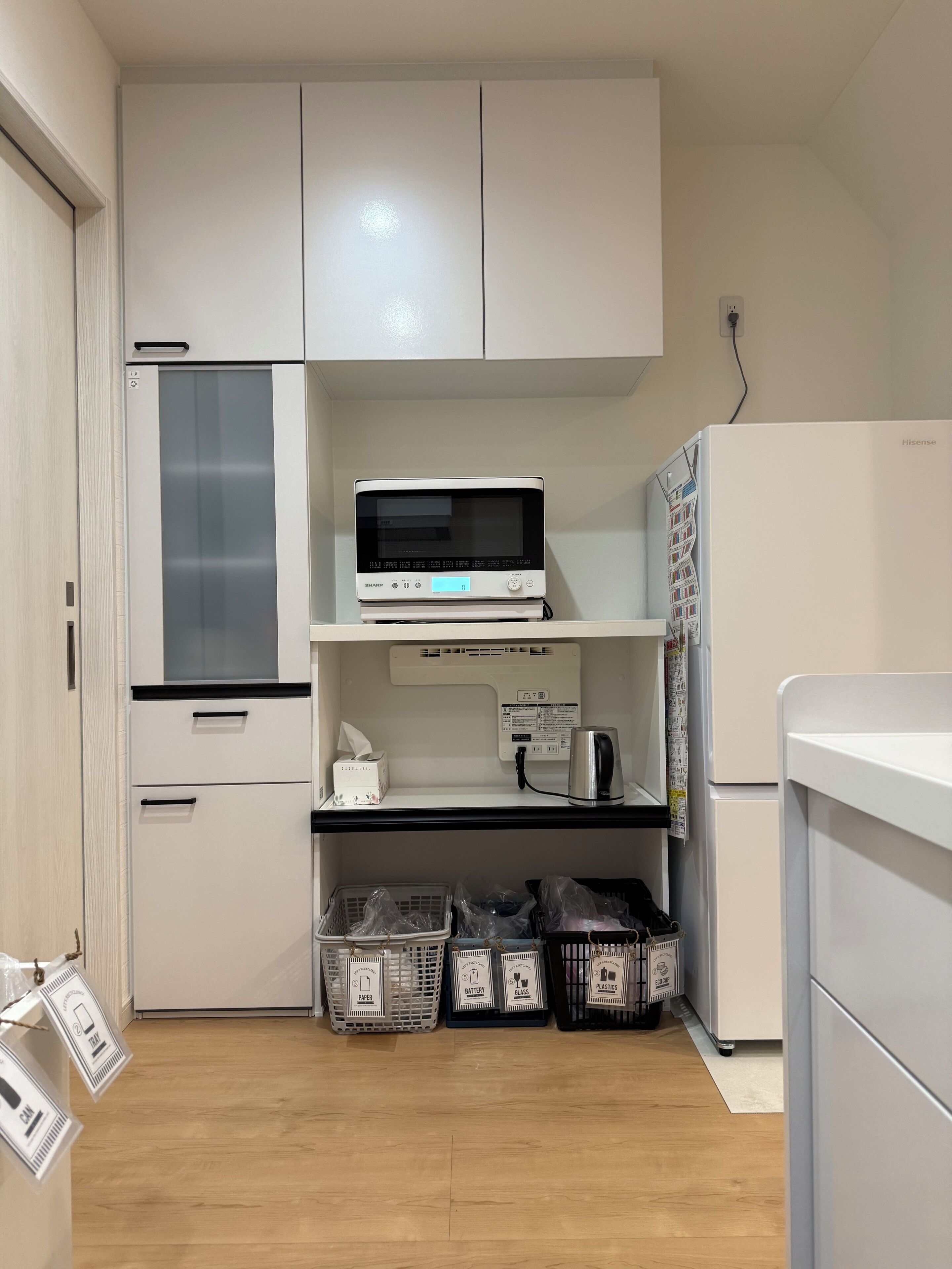 Een compacte keuken met witte kasten, een magnetron, een kleine koelkast en gemarkeerde afvalbakken.