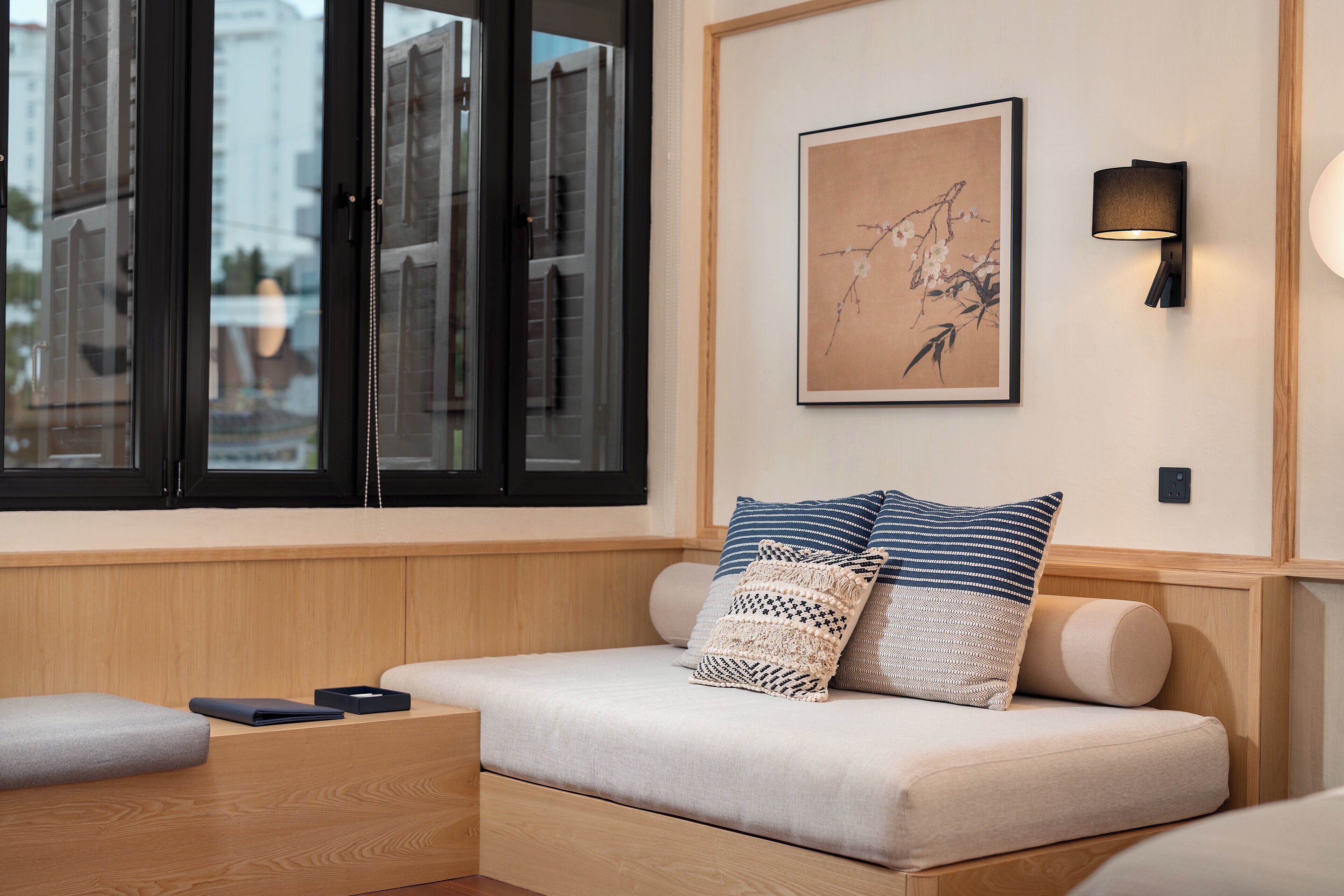 A modern hotel room with a wooden bench, a cushioned seating area, a framed artwork on the wall, and a wall-mounted lamp.