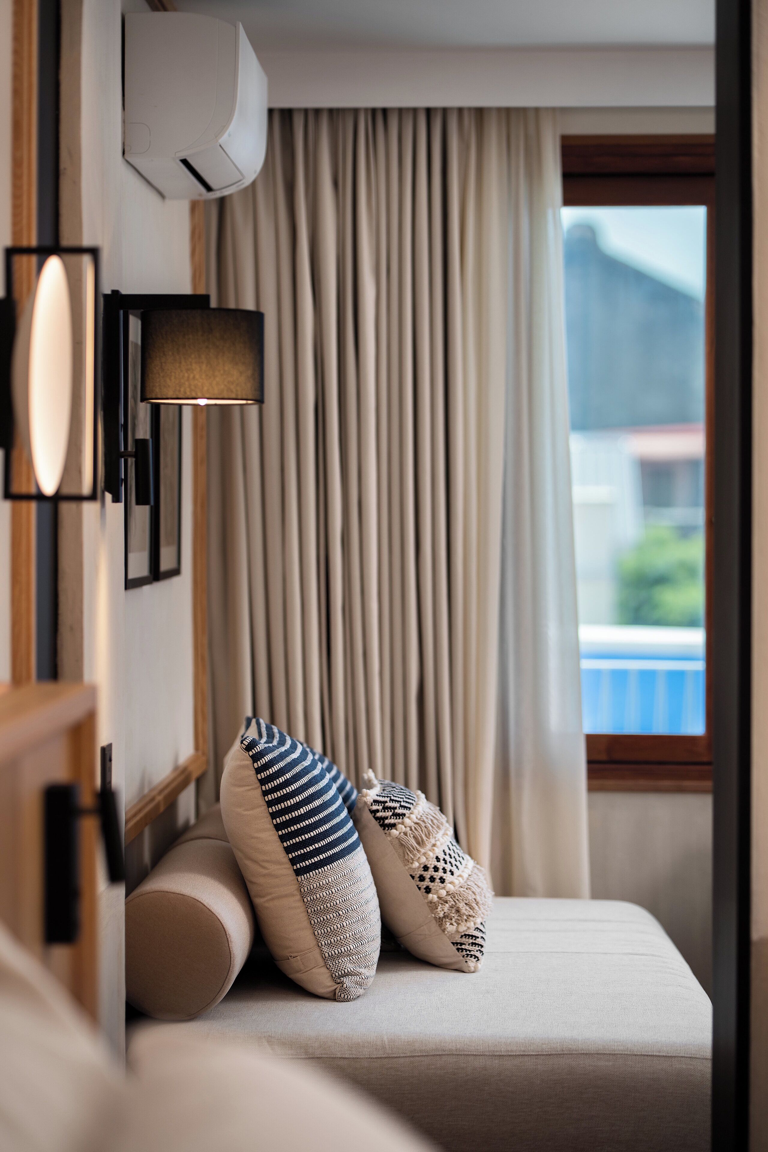 A neatly arranged hotel room with a bed, a wall-mounted air conditioner, a lamp, and a window with curtains.