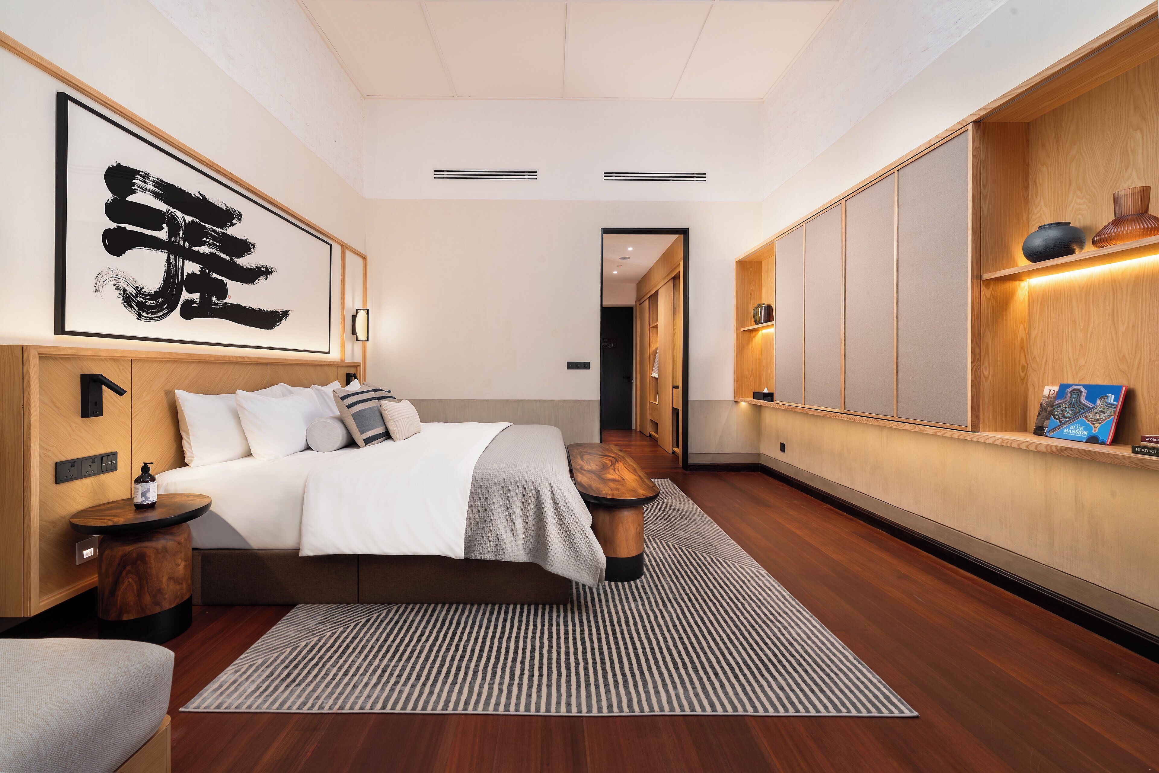 Une chambre d’hôtel moderne avec un grand lit, une tête de lit en bois, une table de chevet, une petite commode, une grande œuvre d’art, un meuble en bois et une petite étagère avec des objets décoratifs.