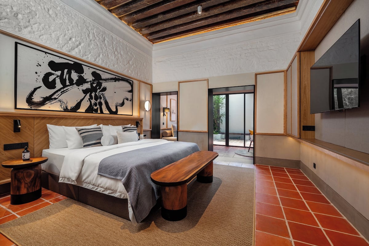 A modern bedroom with a large bed, a wooden bench, a large abstract painting, and a TV mounted on the wall.